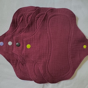 May include: A set of four maroon-colored reusable cloth pads with a textured fabric surface. Each pad features a distinctively shaped absorbent core and a single plastic snap button on the side, with snap colors including light purple, white, black, and yellow.