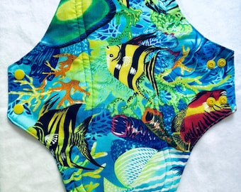 May include: A reusable cloth menstrual pad featuring a vibrant underwater coral reef print with yellow and black angelfish, orange coral, and blue aquatic tones. The pad has a contoured shape with two yellow plastic snap buttons on each side wing.