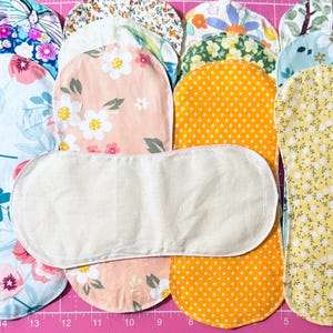 May include: A collection of oblong fabric burp cloths featuring various floral and polka dot patterns in shades of pink, yellow, blue, and green, with one plain cream-colored cloth placed horizontally across the center of the group.
