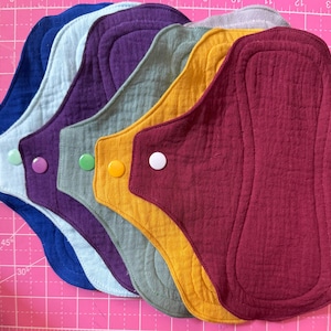 May include: A set of six reusable cloth menstrual pads in various colors including dark blue, light blue, purple, sage green, mustard yellow, and maroon. Each pad features a matching plastic snap button closure and a textured, quilted fabric surface.