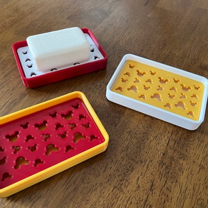 May include: Three soap dishes with a Mickey Mouse design. The dishes are red, yellow, and white. One dish has a white bar of soap inside.