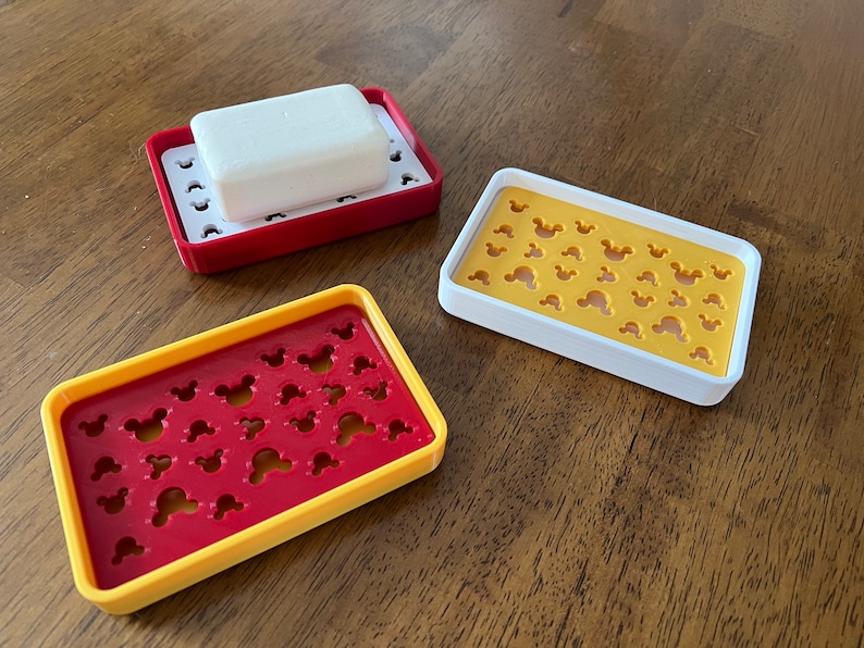 May include: Three soap dishes with a Mickey Mouse design. The dishes are red, yellow, and white. One dish has a white bar of soap inside.