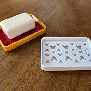 May include: Two white soap dishes with a Mickey Mouse design. The dish on the left has a bar of soap on top. The dish on the right is empty.