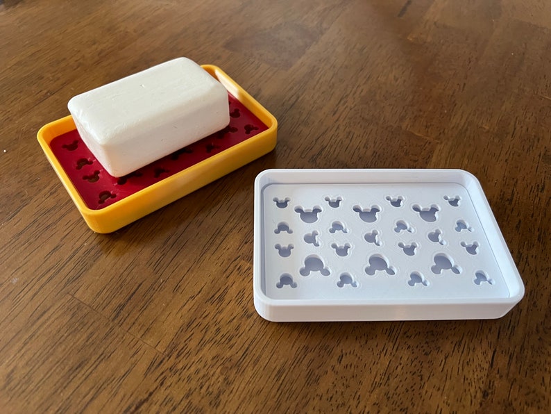 May include: Two white soap dishes with a Mickey Mouse design. The dish on the left has a bar of soap on top. The dish on the right is empty.