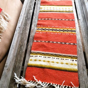 May include: A rectangular, woven textile in shades of red, white, yellow, and brown. The textile features horizontal stripes and geometric patterns. White fringe adorns the bottom edge. The textile is displayed on a weathered wooden tray.