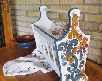 Op de afbeelding: Witte houten tijdschriftenrek met een decoratief bloemmotief in blauw, goud en rood. De rek heeft een handvat en een lattenfront. Het woord "SAMLER" is zichtbaar op een tijdschrift in de rek.