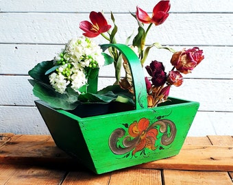 May include: A green wooden basket with a handle, decorated with a floral design. The basket contains white and red flowers and green foliage. The basket is sitting on a wooden surface.