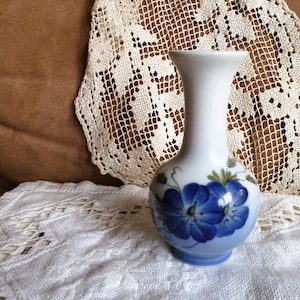 May include: A white ceramic vase with a blue floral design. The vase has a rounded body and a flared neck. The flowers are a vibrant blue with green leaves. The vase is approximately 6 inches tall.
