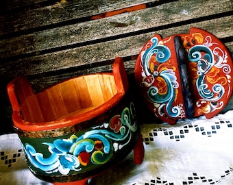 Op de afbeelding: Een ronde houten container met bijpassend deksel, voorzien van een donkergroene buitenband beschilderd met blauwe, witte en oranje bloemmotieven. De container heeft twee opstaande handvatten en een rode voet, met de naam SERA op het deksel.
