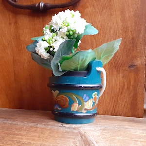May include: A teal-colored wooden bucket-shaped planter with a black trim and floral design, holding white flowers and green leaves. A white rope handle is attached. The planter is on a wooden surface.