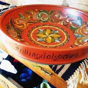 Può includere: Una ciotola ovale in legno rosso con intricati disegni floreali gialli, verdi e marroni. Il bordo presenta il testo "Hallingdalsprøven" in scrittura nera, mostrando uno stile artistico popolare tradizionale.
