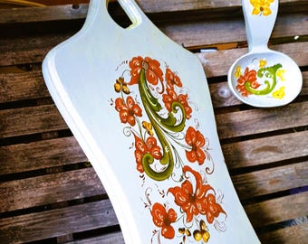 Op de afbeelding: Een lichtblauwe houten snijplank met een handvatgat en een bijpassende lepelhouder, beide met de hand beschilderd met oranje bloemmotieven en groene krullende ranken. De snijplank heeft een gebogen vorm en de lepelhouder heeft een gele bloem.