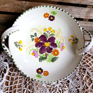 May include: White ceramic serving platter with a floral design. The platter features a central arrangement of purple, orange, and pink flowers with yellow accents. Green trim and handles complete the design. The platter is approximately 12 inches in diameter.