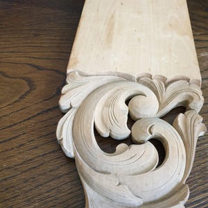 May include: A light-colored wooden corbel featuring an intricate, hand-carved acanthus leaf scroll design. The upper portion consists of a flat, rectangular mounting surface, while the lower section displays detailed, deep-relief floral carvings.