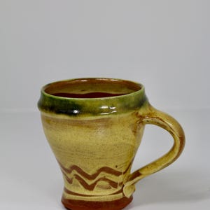 May include: A ceramic mug with a flared rim, featuring a yellow and tan speckled glaze, a green-tinted upper rim, and a brown zigzag pattern near the base. The mug has a curved handle and a solid brown base, showcasing a hand-thrown appearance.