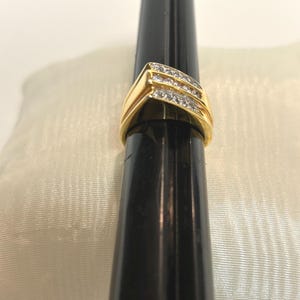 May include: A gold ring with three rows of small, clear stones set in a diagonal pattern. The ring has a thick, polished gold band. The ring is displayed on a black cylindrical object against a cream-colored background.
