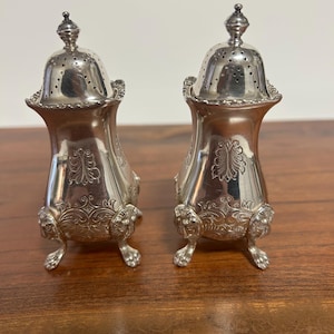 May include: Two ornate silver salt and pepper shakers. Each shaker has a perforated lid, a flared body with engraved floral designs, and rests on three claw feet. The shakers are displayed on a wooden surface.