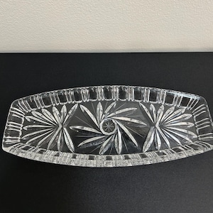 May include: A clear, elongated glass tray featuring a decorative cut pattern. The design includes three distinct starburst motifs across the center and a series of vertical rectangular indentations along the raised, scalloped edges of the dish.