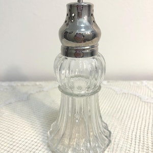 May include: A clear glass sugar shaker featuring a vertical ribbed pattern on the body and a polished silver-tone metal lid with a small finial at the top and multiple dispensing holes around the upper section of the cap.