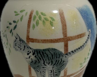 May include: A cream-colored ceramic vase featuring a painted illustration of a grey tabby cat standing on a brown window ledge. The design includes a brown window frame, green leaves, a yellow sun-like shape, and a blue sky area.