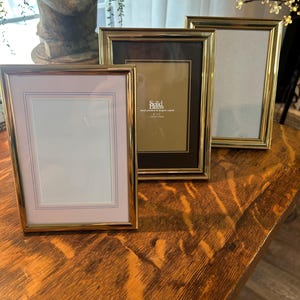 May include: Three rectangular picture frames with polished gold-tone metal borders. The center frame contains a dark insert with the text Solid Brass, hand polished & lacquer coated, 5 x 7, 13cm x 18cm. Each frame features a light-colored matting border.