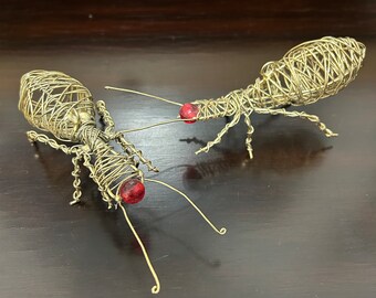 May include: Two gold wire sculptures of ants with red glass beads for eyes. The ants are posed as if they are facing each other.