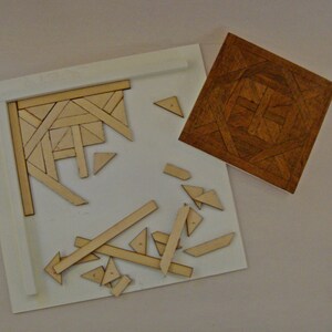 May include: A wooden puzzle with a partially assembled section and scattered pieces. The puzzle features a geometric design with various shapes and angles. A separate, completed square wooden piece with a patterned design is also present.
