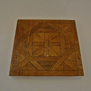 May include: Square wooden decorative tile with a geometric pattern. The tile features a repeating design of interlocking shapes and lines in shades of brown. The edges of the tile are a lighter color, creating a border.