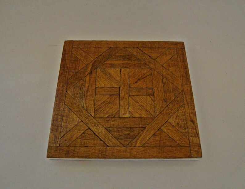May include: Square wooden decorative tile with a geometric pattern. The tile features a repeating design of interlocking shapes and lines in shades of brown. The edges of the tile are a lighter color, creating a border.