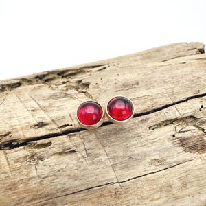 May include: A pair of stud earrings featuring round, deep red cabochon stones set in silver-colored metal. The earrings are displayed on a weathered, light brown wooden surface, creating a contrast between the polished jewelry and the rustic background.