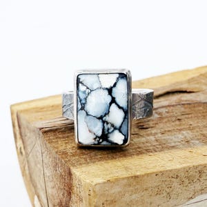 May include: A rectangular ring with a white, blue, and black stone set in a silver band. The stone has a unique, marbled pattern. The ring is displayed on a wooden surface, showcasing its design and craftsmanship. A piece of jewelry.