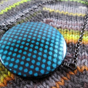 May include: A teal blue pendant necklace with a black chain. The pendant is round and has a pattern of small teal blue dots on a black background.