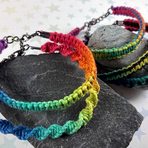 May include: Three rainbow colored crocheted bracelets with metal clasps. The bracelets are layered on top of each other on a gray rock.