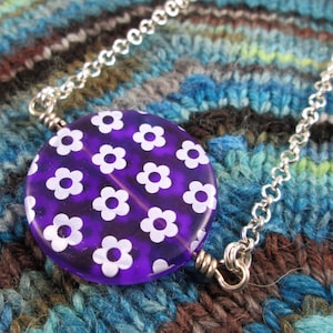 May include: A silver chain necklace with a purple round pendant featuring a white floral pattern.