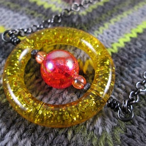 May include: A yellow, glittery, round pendant with a red, iridescent glass bead in the center. The pendant is attached to a black chain.
