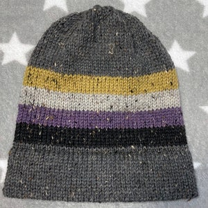 May include: A knit beanie featuring a speckled texture throughout. The design includes a large dark gray base, followed by horizontal stripes in mustard yellow, light gray, lavender, and black, finished with a wide dark gray ribbed cuff.