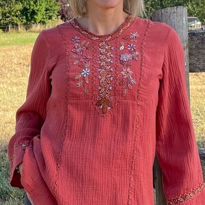 May include: A woman wearing a long-sleeved, rust-colored blouse with embroidered floral details. The blouse has a round neckline and a relaxed fit. She is wearing blue jeans and has a casual, natural look.