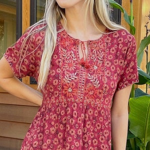 May include: A red midi dress with a floral print and embroidered neckline. The dress has short sleeves and a tie at the neckline.