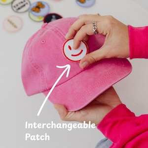 May include: A pink baseball cap with an interchangeable patch featuring a smiling face. Additional patches with various designs and text are scattered around. The image shows the patch being applied to the cap. The text in the image says "Interchangeable Patch".