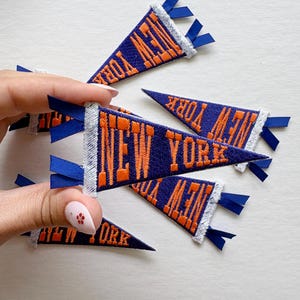 May include: A collection of small, triangular pennant-shaped patches featuring the text NEW YORK embroidered in orange thread on a dark blue fabric background. Each patch has a white stitched border and small blue ribbon tabs at the base.