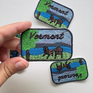 May include: Rectangular embroidered patches featuring a landscape scene with green grass, a blue lake, mountains, and two brown chairs. The word Vermont is stitched in dark brown thread across the top of each patch with a dark gray border.