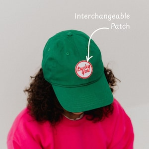 May include: A green baseball cap with a curved brim. The cap features a circular patch with the words "Lucky You" in red script. The patch is outlined in red and set against a white background. The cap is made of a soft material.