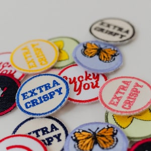 May include: A selection of embroidered patches with diverse designs and text. Some patches display "EXTRA CRISPY" in various colours, while others feature a butterfly or the phrase "lucky you". The patches are circular and have a stitched border.