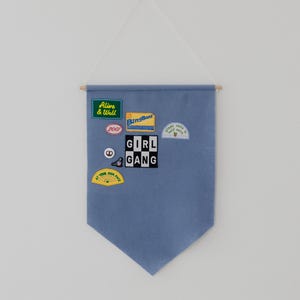 May include: A blue felt banner with various embroidered patches. Patches include text that reads "Alive & Well", "BingBong", "Girl Gang", and "At Your Own Pace". The banner is suspended by a wooden dowel and string.