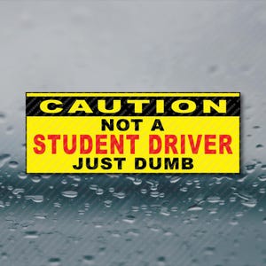 May include: A rectangular sticker with a yellow background and black and red text. The text reads "CAUTION NOT A STUDENT DRIVER JUST DUMB". The sticker is designed to be placed on a vehicle.