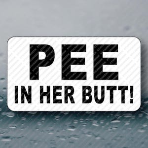 May include: A rectangular sticker with rounded corners, featuring the text "PEE IN HER BUTT!" in bold, black letters. The sticker has a white background and is set against a blurred, possibly wet, surface.