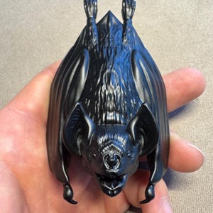 May include: A matte black figurine of a bat in a hanging position with its wings folded against its body. The bat features detailed fur texture on its back, pointed ears, and a small snout with an open mouth, showcasing a realistic anatomical design.