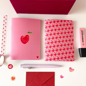 May include: A collection of pink stationery items, including two notebooks, one with a cherry illustration and the other with a heart pattern. Also included are a red box, pen, stickers, washi tape, and a highlighter.