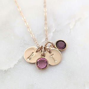 May include: Gold-colored necklace with three small, round pendants. Two pendants have the letters "B" and "T" engraved on them. The necklace also features two purple gemstones. The chain is delicate and the background is white.
