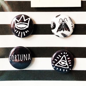 May include: Four black and white witchy themed buttons with white designs on a black background. The buttons are arranged in two rows of two. The top row features a crown with an eye and a moth with wings. The bottom row features the word "Fortuna" and a triangle with an eye.